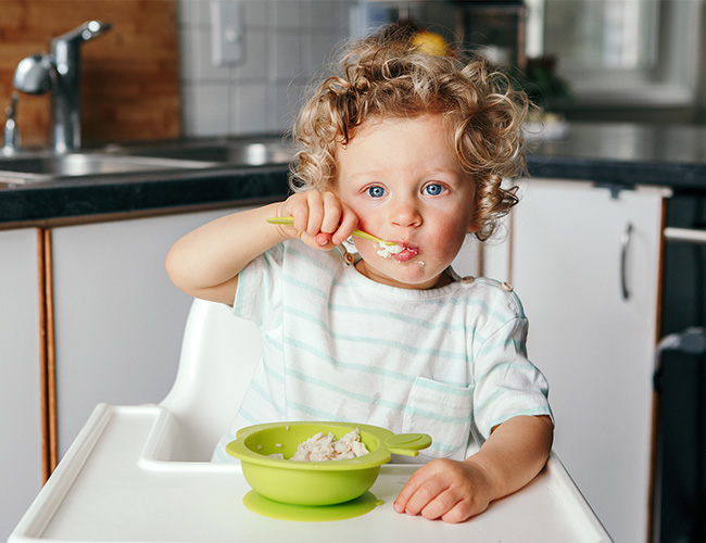 12 - 18 month old kid independently eating soft cereal with a spoon.