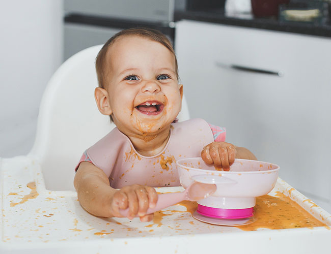 6 - 9 month old kid eating with hands and a spoon.